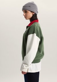 Zelenobílá mikina s červeným límcem, s volným střihem a dlouhými rukávy, doplněná šedou pletenou čepicí.