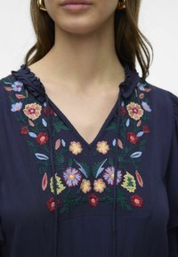Mornarica bluza s cvjetnim vezom u crvenoj, ružičastoj, plavoj i žutoj boji. Ima V-izrez i nabran rub na ovratniku. Teksturirana tkanina.