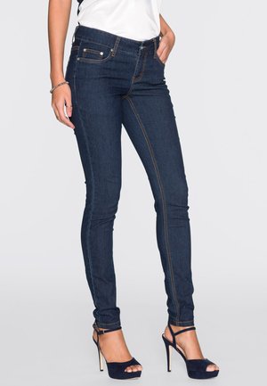 Jean skinny en denim noir avec des coutures orange contrastantes, cinq poches et une taille mi-haute, associés à des sandales à talons noires.