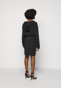 Žena s afro účesem, oblečená v černé mikině s kapucí, černé sukni s bílými postranními pruhy a černých sandálech na vysokém podpatku, otočená zády.