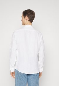 Camisa de lino blanca de manga larga con cuello mandarín, detalles de costura visibles y dobladillo curvado, combinada con jeans de color azul claro.