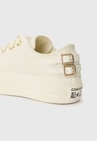Kremasti platneni tenisice s zlatnim rupicama, dvostrukim kopčama sa strane i bijelim gumama koje imaju logo "Converse All Star".