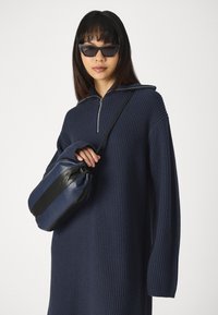 Marineblauwe geribbelde gebreide jurk met halve rits kraag. Individu houdt een marineblauwe en zwarte gestreepte handtas met een gladde textuur vast. Donkere zonnebril gedragen.