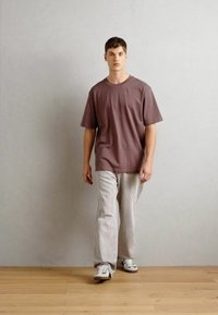 T-shirt oversized em mauve combinado com calças largas em cinza claro. O modelo usa ténis com detalhes em verde. Texturas de tecido suaves e casuais.