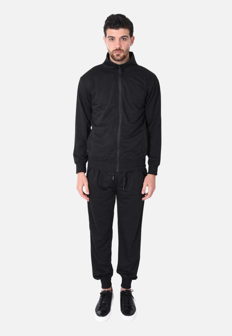 Giacca nera con zip e colletto alto, abbinata a pantaloni tuta neri. Tessuto liscio, polsini a costine e vita con cordino. Indossato con sneakers nere.