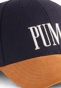 Gorra marina con visera de ante color marrón claro, que presenta el logotipo "PUMA" bordado en blanco en la parte frontal. Textura de tela suave con detalles de costura.