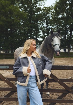 Grå shearlingjakke med en hvid pelskrave og cuffs, parret med lyseblå jeans. En grå hest står i baggrunden nær et træhegn.
