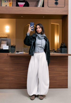 Tânără purtând o geacă de piele neagră, hanorac gri și pantaloni largi albi făcând un selfie într-o cameră modernă cu panouri din lemn.