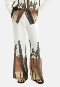 Lnianie outfit składający się z koszuli z guzikiem i spodni o szerokich nogawkach. Biała baza z zielonymi kaktusami i brązowym krajobrazem pustynnym. Płaskie czarne sandały.