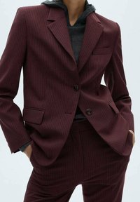 Personne portant un costume à rayures bordeaux avec un blazer à double boutonnière et un pantalon assorti sur un sweat à capuche gris foncé, les mains dans les poches.