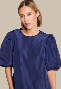 Marineblå puffermetopp, glatt satengstoff, rund halslinje, rynker på ermene, avslappet passform. Passer til uformelle eller semi-formelle anledninger.