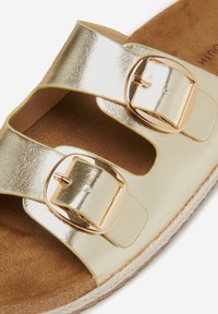 Gouden metalen sandalen met twee brede banden, voorzien van gouden gespen en een natuurlijke tan voetbed, tegen een gestructureerde zool.