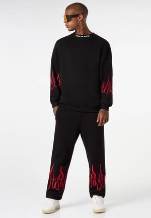 Completo di felpa nera e pantaloni con grafiche di fiamme rosse sulle maniche e sui orli dei pantaloni. Il materiale sembra morbido con una vestibilità rilassata. Sneakers bianche.