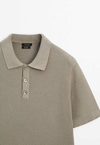 Beige lyhythihainen poolopaita teksturoidulla neuloksella. Siinä on tasainen kaulus ja neljä hopeanväristä nappia pliseerauksessa.