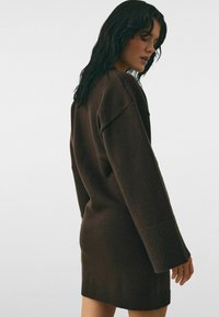 Frau mit nassen dunklen Haaren, die ein langes, braunes Strickkleid mit gerippten Bündchen und Saum trägt, stehend vor einem schlichten hellen Hintergrund.