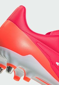 Scarpino da calcio rosa brillante con una parte superiore in pelle testurizzata, caratterizzato da un tallone arancione a contrasto e una suola bianca con tacchetti metallici per la trazione.