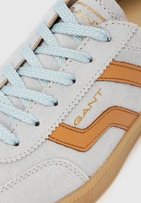 Hellgrauer Wildledersneaker mit hellblauen Schnürsenkeln, braunen Lederstreifen, hellbrauter Gummisohle und "GANT"-Logo in goldener Schrift.