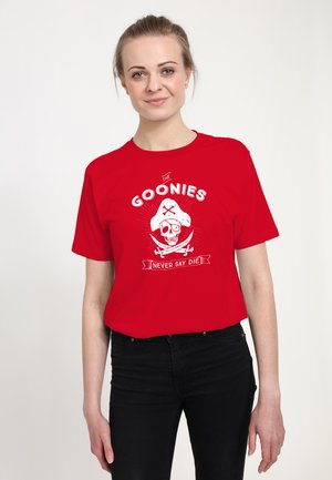 Ung kvinde med lyst hår sat tilbage iført en rød T-shirt med hvidt piratskalle og krydsknogler samt teksten "The Goonies Never Say Die".