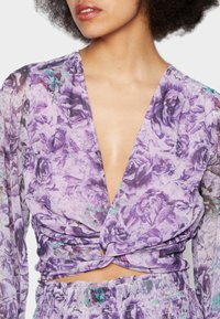 Top floral roxo com mangas longas, apresentando um decote V profundo, detalhe torcido na cintura e uma textura leve e transparente.