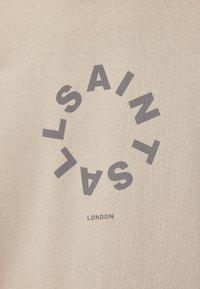 Beige pamut póló, sötétszürke kör alakú grafikával, középen a "SAINT" és "LONDON" felirattal.