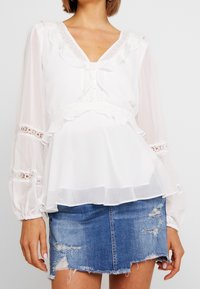 Weiße Bluse mit gerafftem Ausschnitt und langen, transparenten Ärmeln, die Spitzenakzente aufweist, kombiniert mit einem zerstörten Jeansrock.