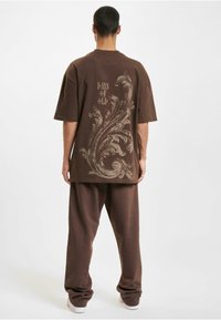 Brun oversized t-shirt med dekorativt lysebrunt svirlemønster og teksten "DAYS OF OLD" på ryggen, parret med matchende afslappede bukser.