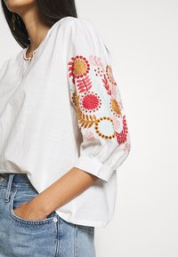 Blusa di lino bianca con maniche a palloncino, caratterizzata da un ricamo floreale colorato in rosa, rosso e oro. Vestibilità casual con scollatura rotonda.