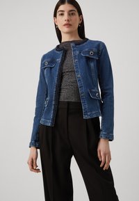 Kvinnelijk in een blauwe spijkerjack over een grijze geribde top en losse zwarte broek, staand tegen een effen lichte achtergrond.