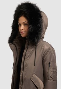 Ruskea toppatakki, jossa mustalla karvareunuksella varustettu huppu, vetoketju ja painonapit edessä, sivutaskut sekä vetoketjullinen hihatasku.