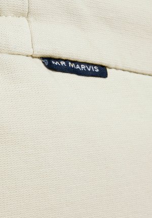 Gros plan sur un vêtement en maille couleur crème avec une petite étiquette en tissu noir portant l'inscription "MR MARVIS" cousue près du col.