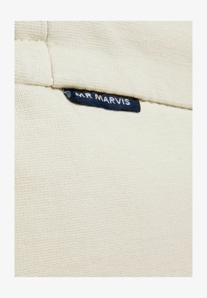 Gros plan sur un vêtement en maille couleur crème avec une petite étiquette en tissu noir portant l'inscription "MR MARVIS" cousue près du col.