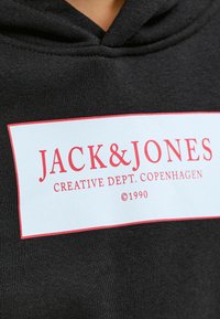 Sudadera negra con una etiqueta blanca con texto rojo que dice "JACK&JONES CREATIVE DEPT. COPENHAGUE ©1990". Textura de tela suave.
