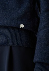 Tmavomodrý vlnený sveter s rebrovaným lemom a manžetami. Obsahuje kovovú značku loga blízko lemu. Mäkká štruktúra s jemnými farebnými fliačkami.