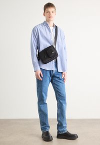 Borsa a tracolla nera con zip, caratterizzata da una tasca frontale, indossata da un modello con una camicia a righe blu e bianche e jeans blu, su uno sfondo neutro.