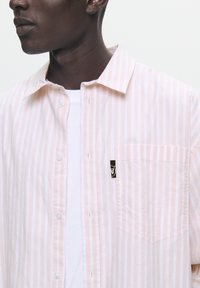 Cămașă button-up cu dungi roz pal și alb, dotată cu un buzunar pe piept și un mic eticheta neagră cu logo, fabricată dintr-un material moale și ușor.