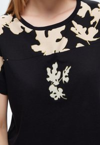 Svart t-shirt med en krämfärgad blommönster på den övre delen, med en mjuk textur och rund halsringning. Mjuk tyg.