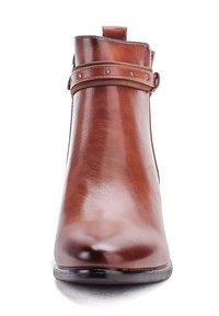 Botín de ante marrón suave con una correa decorativa y pequeños tachuelas metálicas, punta redondeada y suela negra.