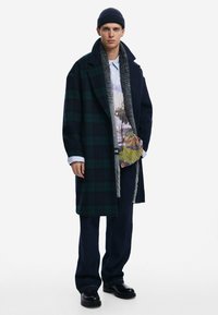 Long navy coat with green plaid design, layered over a patterned shirt, paired with dark blue pants and black boots. Features grey textured scarf.