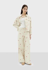Beige denimjack en wijde broek met rode bloemenborduursels. Het model draagt een witte tanktop en beige schoenen, met voorzakken.