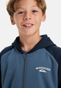 Hoodie met rits in blauw en marineblauw, voorzien van een wit Quiksilver-logo op de borst en raglanmouwen. Zachte stof met een gladde textuur.