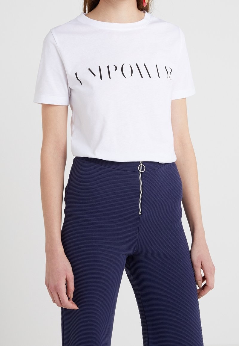 Wit katoenen T-shirt met een zwart, gebogen tekstontwerp dat "EMPOWER" leest. Gecombineerd met ribbenachtige marineblauwe broek met een ritsdetail.