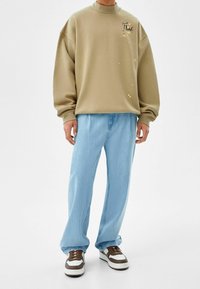 Beige oversized sweatshirt med hög krage och svart textgrafik, i kombination med ljusblå avslappnade jeans och sneakers med bruna accenter.