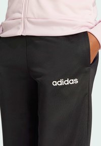 Črne športne hlače iz gladkega blaga, z belim logotipom "adidas". Nabrano pasu dodaja teksturo. Ob strani je vidna roza majica.