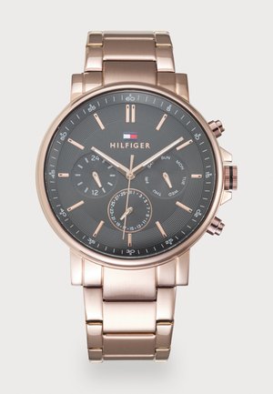 Montre-bracelet en métal rose doré avec un cadran noir, trois sous-cadrans pour l'heure 24 heures, le jour et la date, et le logo Tommy Hilfiger en dessous de 12 heures.