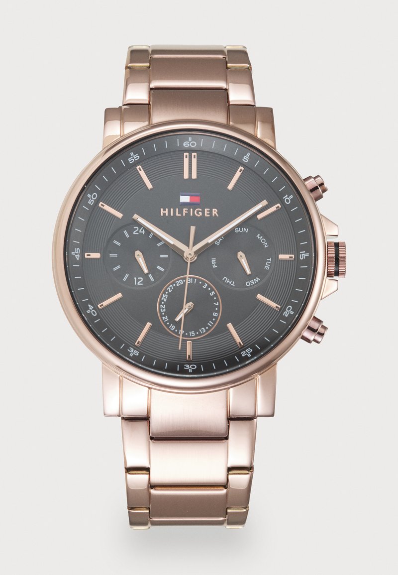 Ροζ χρυσό μεταλλικό ρολόι χεριού με μαύρο καντράν, τρία υποκαντράν για 24ωρη ώρα, ημέρα και ημερομηνία, και το λογότυπο της Tommy Hilfiger κάτω από τις 12 στη θέση του ρολογιού.