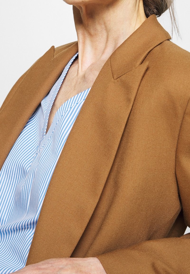 Mujer con un blazer marrón sobre una blusa a rayas azul y blanca, mostrando el cuello, el hombro y parte del brazo frente a un fondo liso.