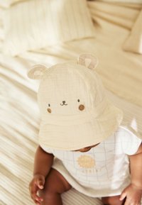 Vedeni klobuk kremaste barve z medvedjimi ušesi, okrašen z vezenimi detajli obraza. Mehkoba teksture tkanine, večplastna zasnova, na nevtralnem ozadju.