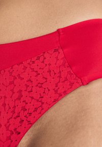 Rote Unterwäsche mit einem glatten oberen Band und einem durchsichtigen Spitzen-Einsatz mit floralem Muster, die eine Mischung aus Texturen und Farben betont.