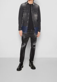 Giubbotto di jeans nero con pannelli azzurri più chiari, due tasche frontali, jeans neri usurati con dettagli strappati e stivali neri lucidi.