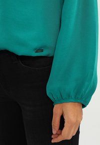 Tyrkis langærmet bluse med samlede manchetter, parret med sorte jeans, hvor en hånd er afslappet ved siden af.